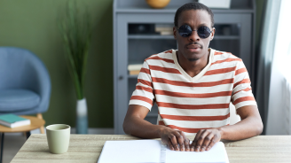 Front view portrait of adult black man reading tactile book in braille and wearing dark sunglasses, accessible education concept